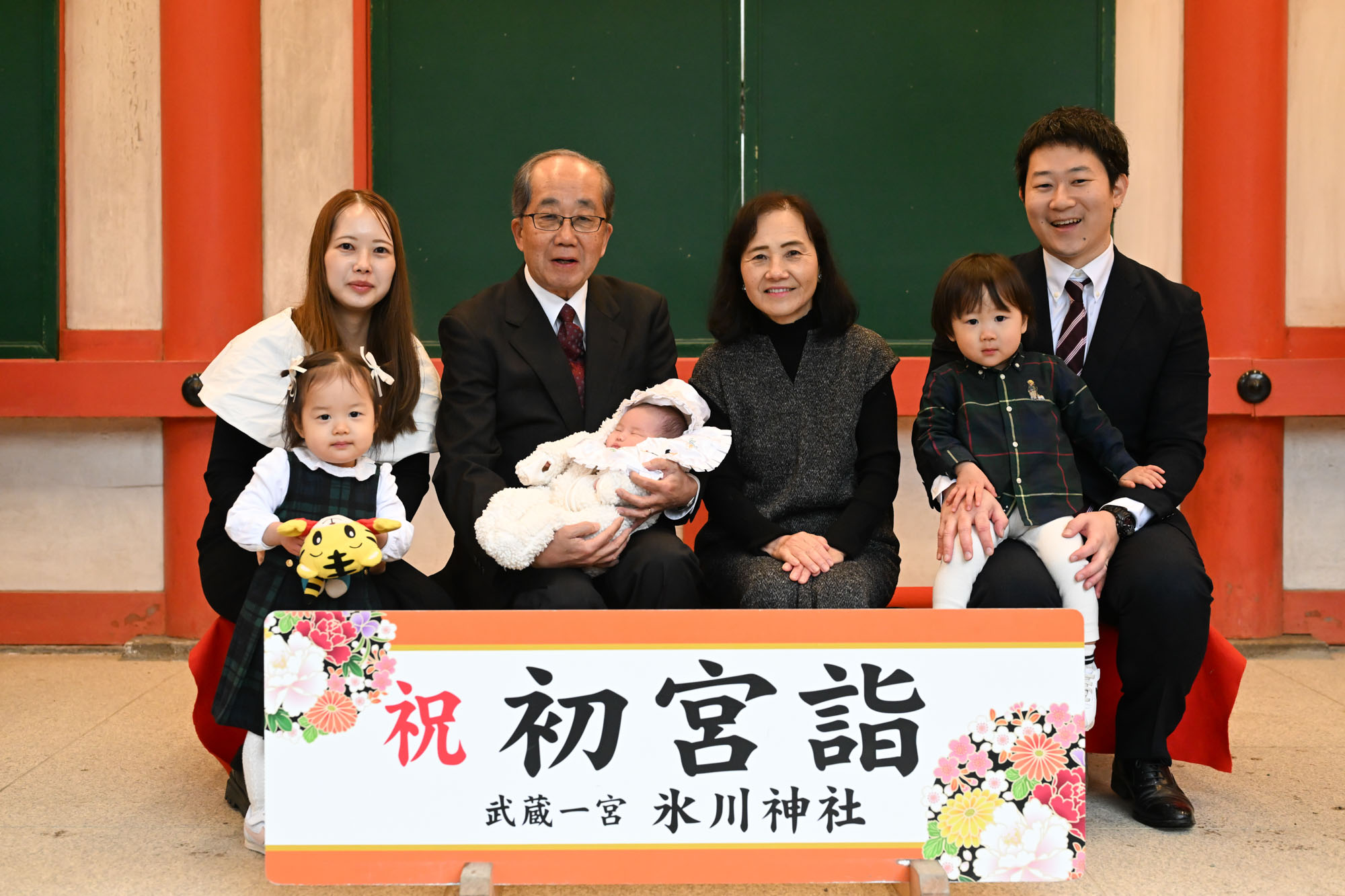 大宮氷川神社の特設看板前で、家族みんなで椅子に座って撮ったお宮参り記念写真。雨の日でもリラックスした雰囲気で残せる貴重な1枚。