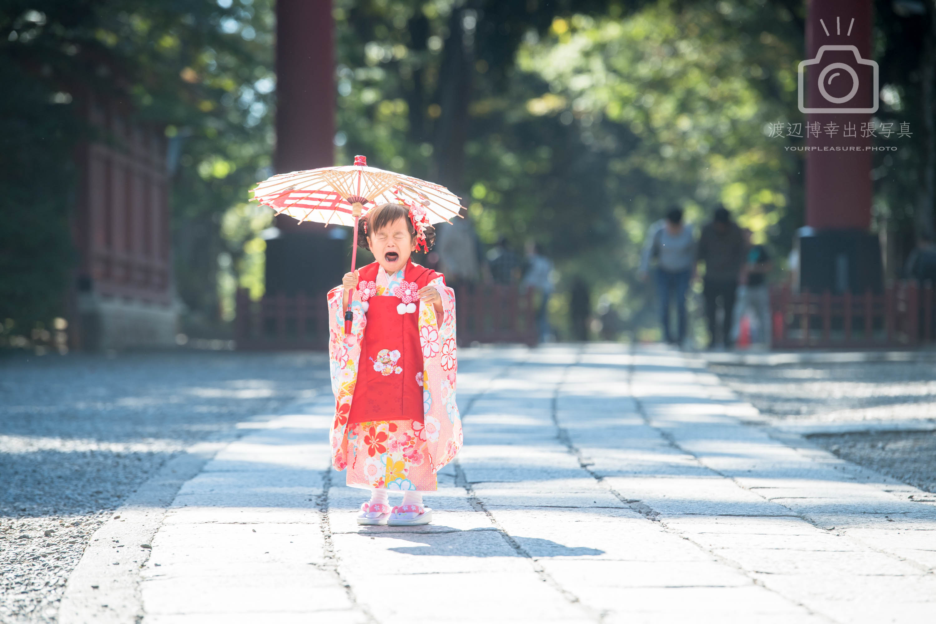 神社の参道で赤い着物を着て和傘をさし立ちながら泣いている女の子