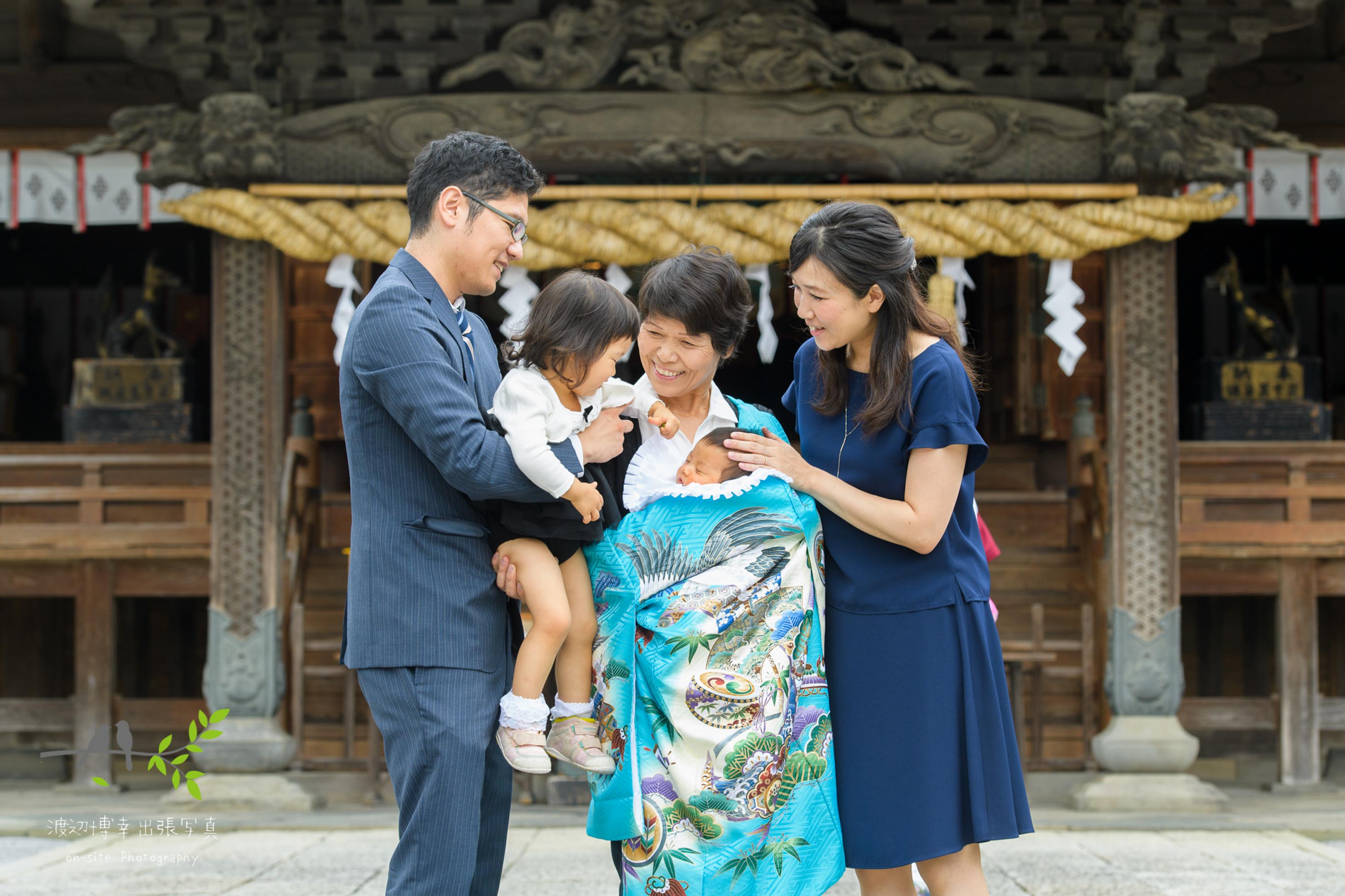 神社本殿前で掛着姿で赤ちゃんを抱く祖母と横に立つ家族