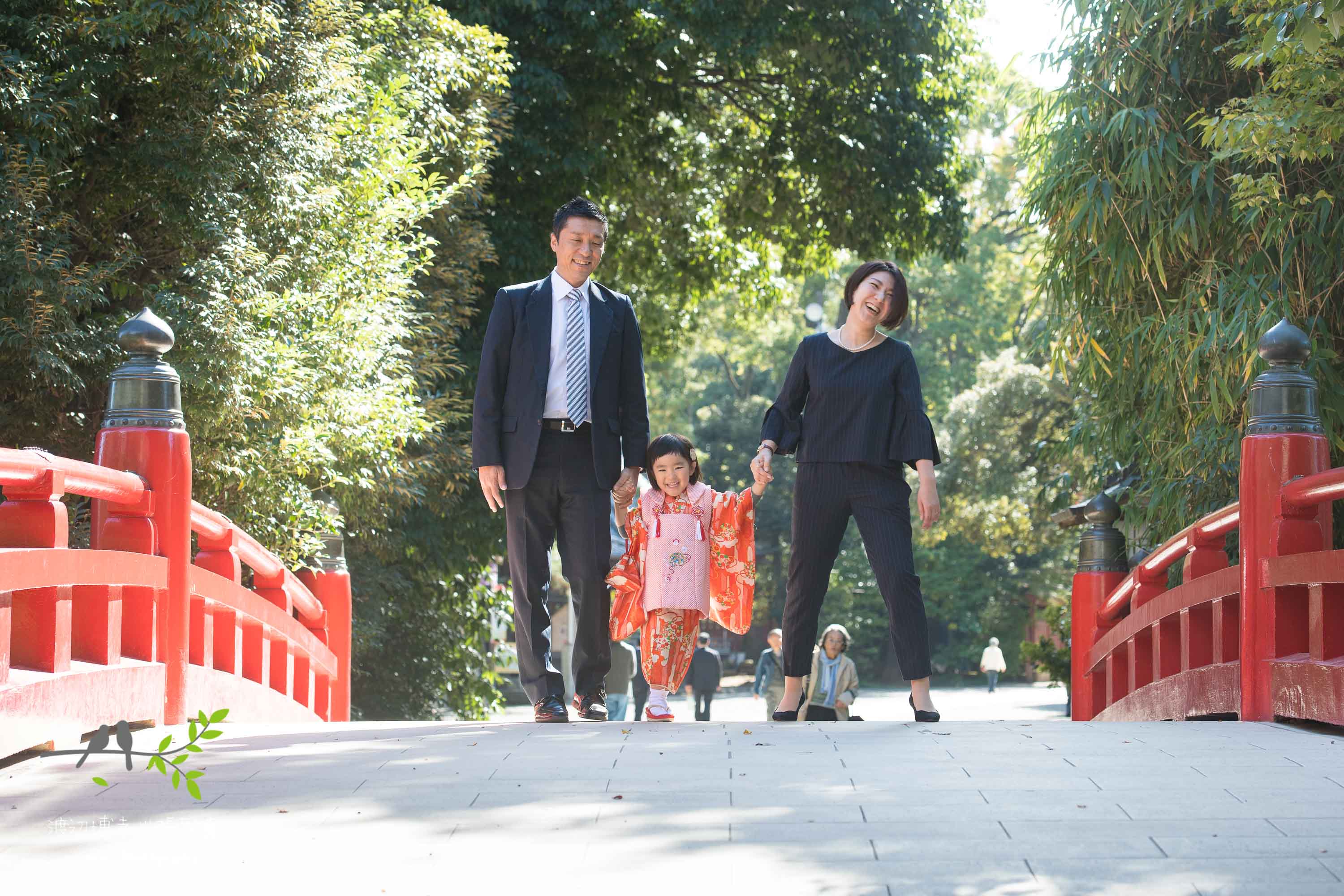 赤い橋の上を歩くオレンジ色の着物姿の小さな女の子とパパとママ