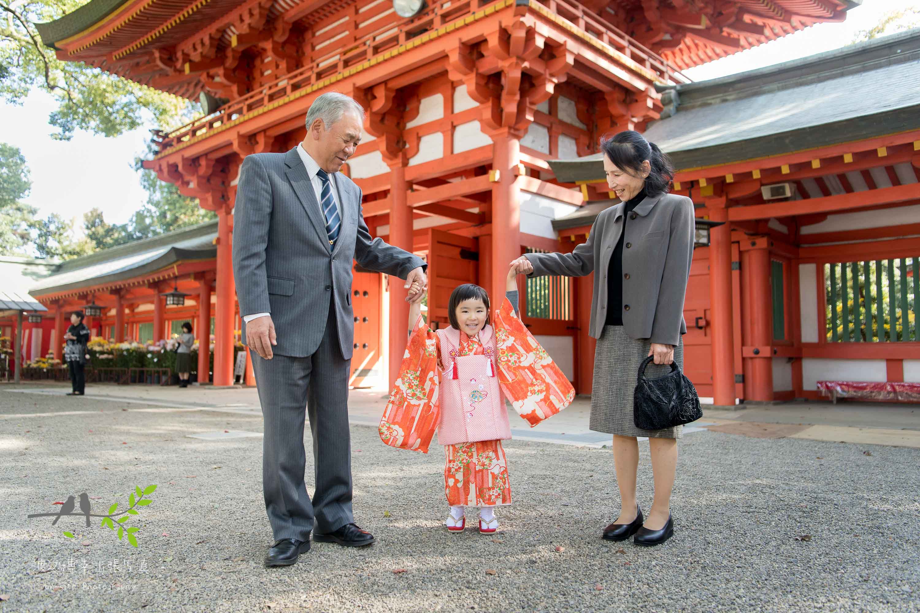 オレンジ色の山門の前で立っているオレンジ色の着物を着た女の子と祖父と祖母