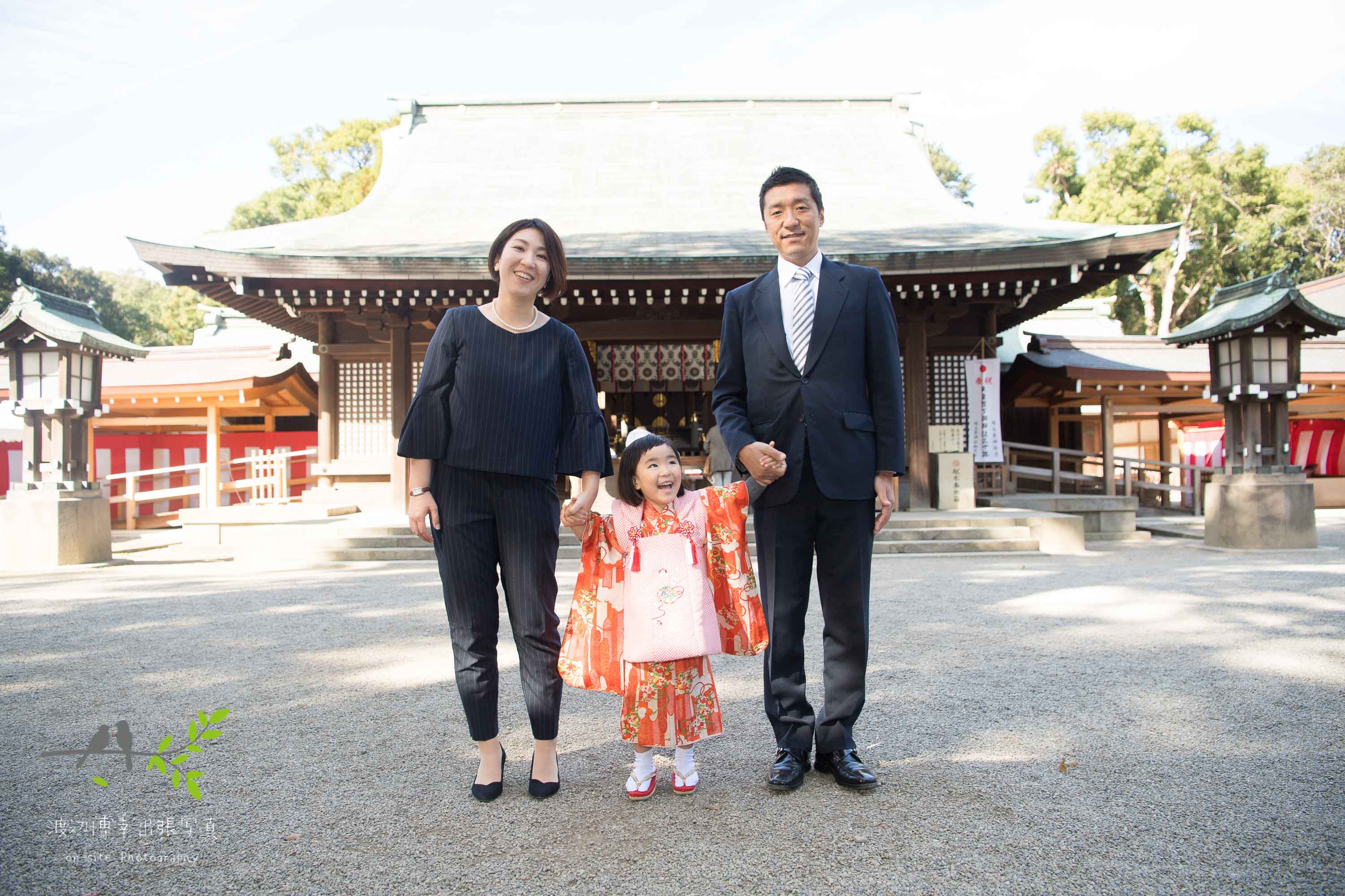 神社本殿の前に並ぶオレンジ色の着物を着た女の子とパパとママ