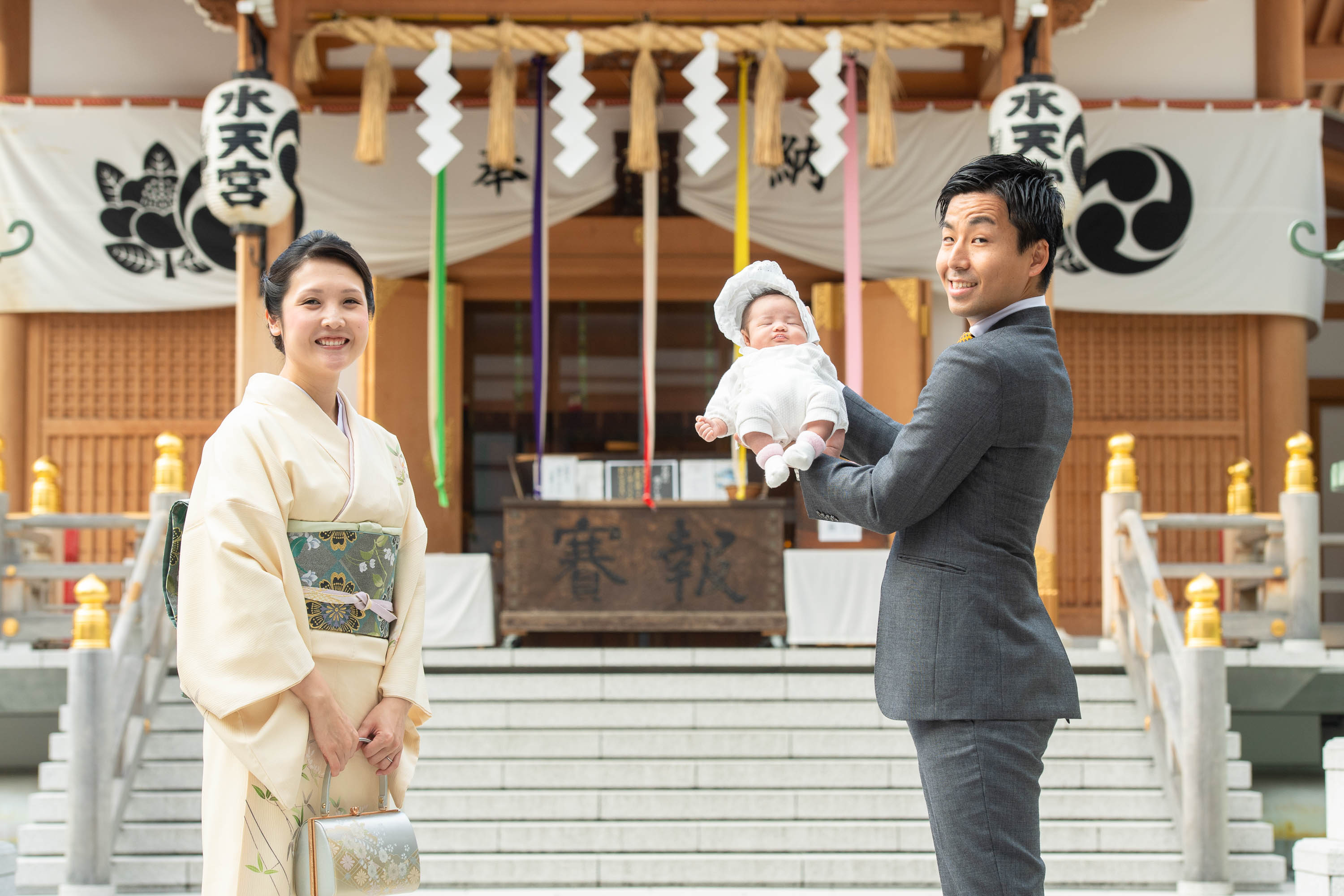 赤ちゃんを手で持ち上げ抱くスーツ姿の男性と横に立つ和装の女性