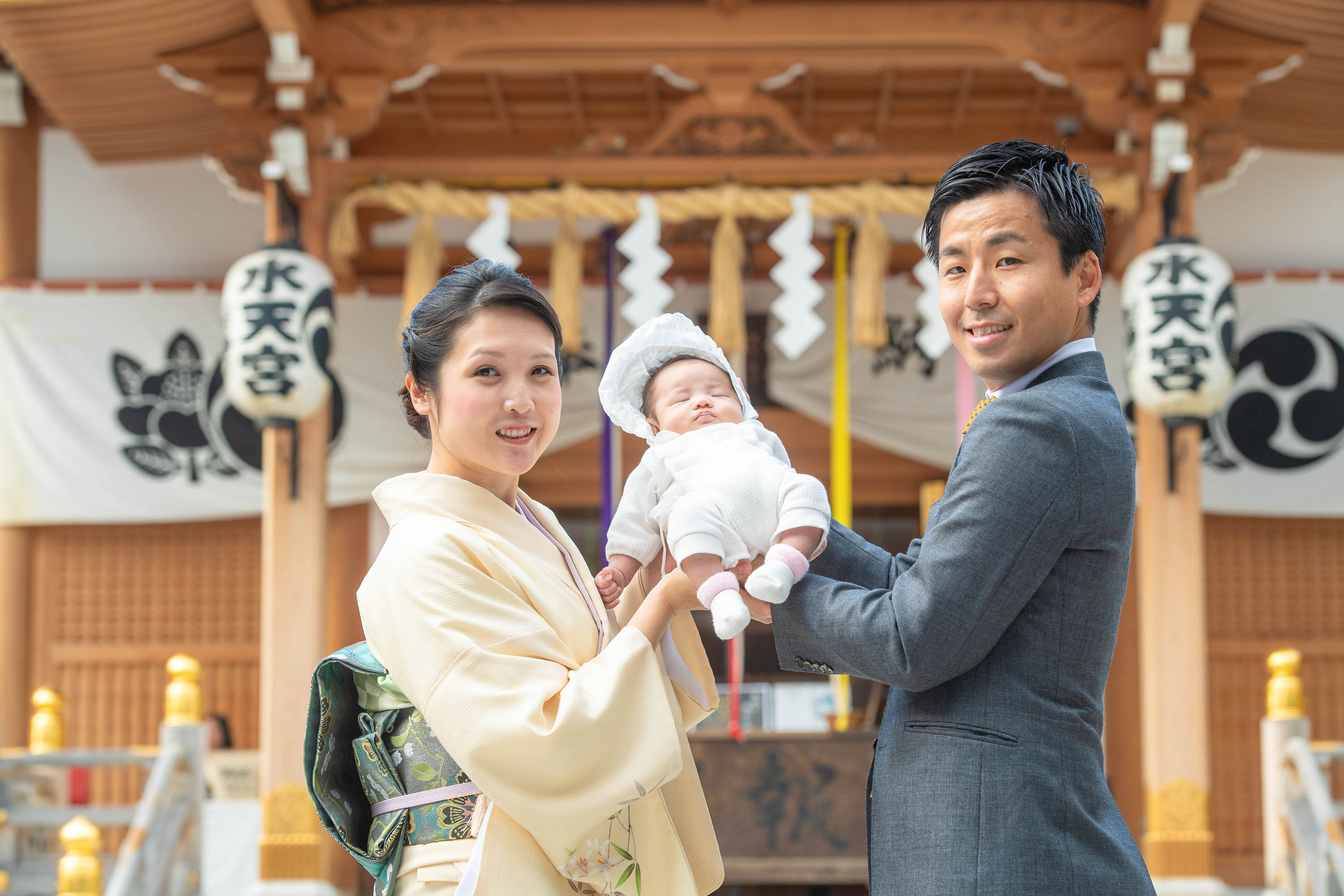 赤ちゃんを手で持ち上げ抱くスーツ姿の男性と横に立つ和装の女性