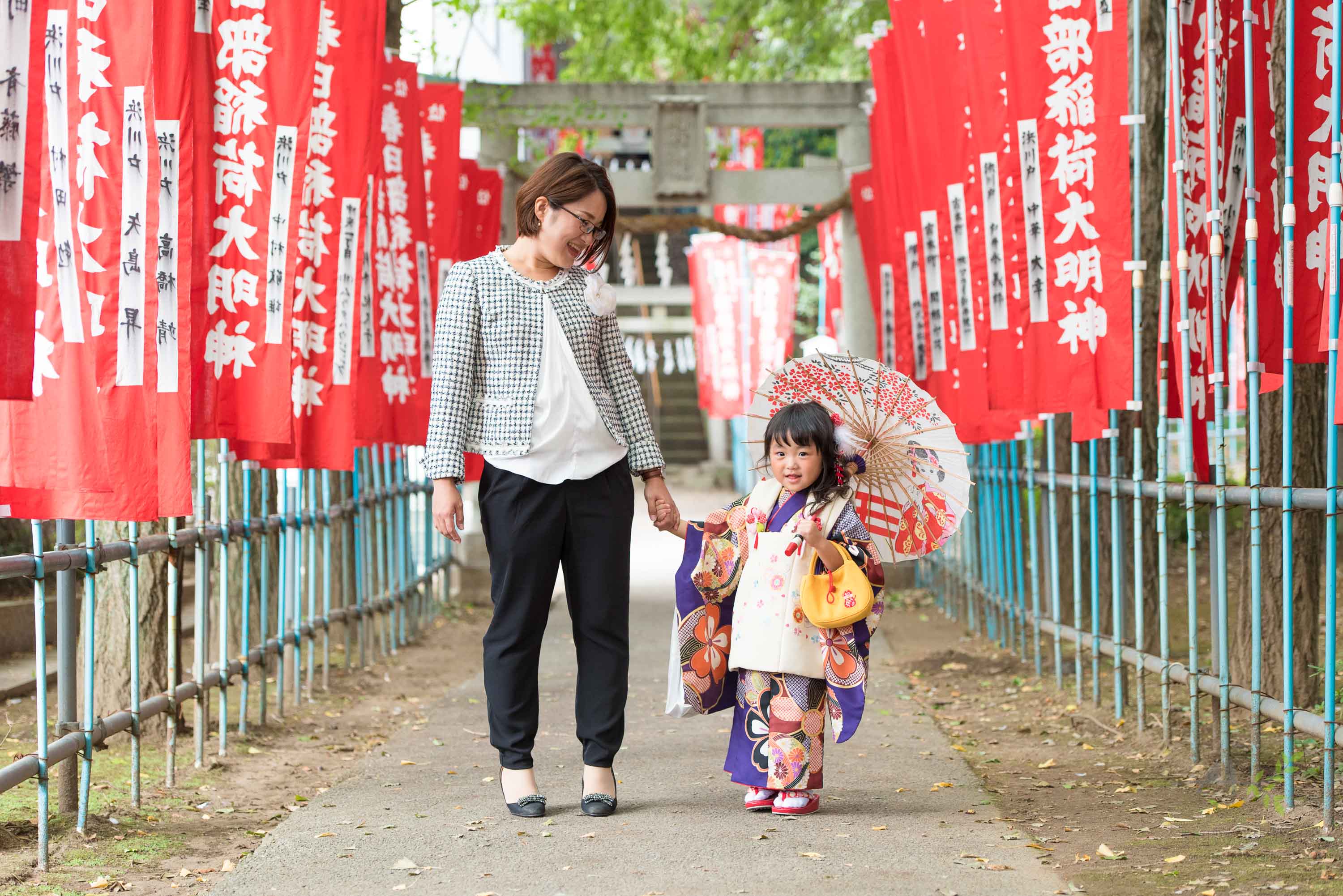 赤い神社のぼりの参道に立つ小さな和傘をさした七五三の着物姿の女の子と女性