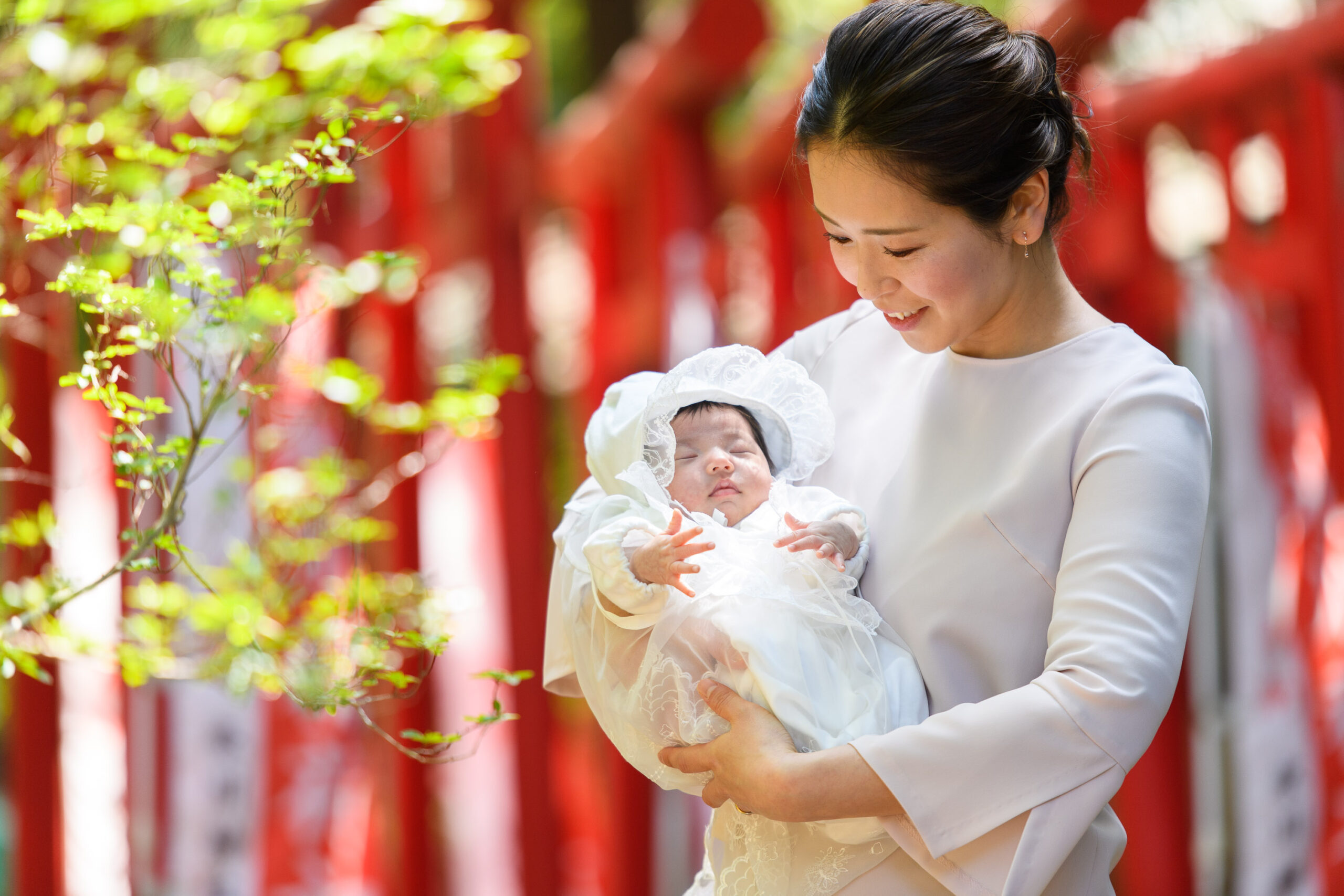 大宮氷川神社の境内に差し込む柔らかな光の中で赤いたくさんの鳥の前で赤ちゃんを抱っこするお宮参りの女性