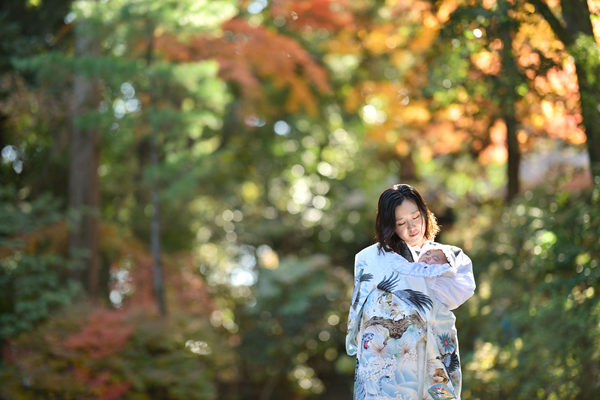 画像 大宮氷川神社のお宮参りで、神社裏手の紅葉の柔らかな光の中で白い掛け着をつけ、赤ちゃんを抱く女性