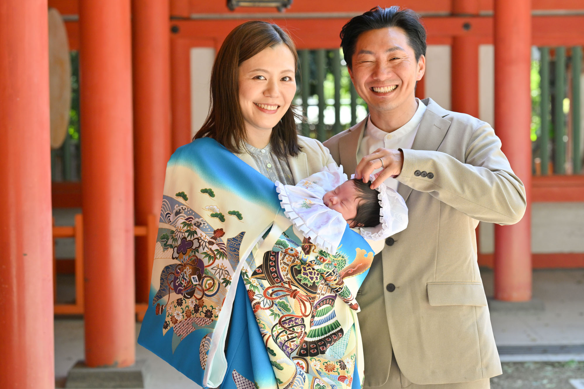 大宮氷川神社のお宮参りで、赤い柱のところで、青い掛け着姿で赤ちゃんを抱っこするママ横に立つパパ