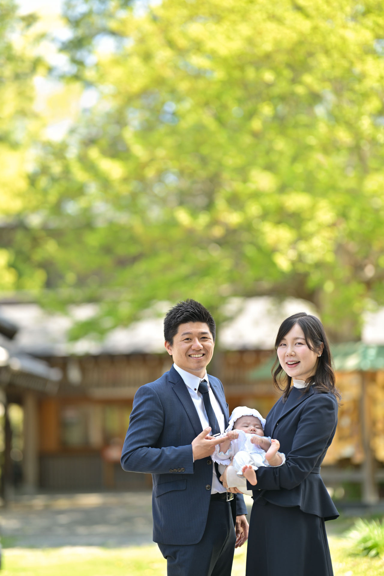 和樂備神社のお宮参りできれいな木をバックで赤ちゃんを抱っこするご夫婦