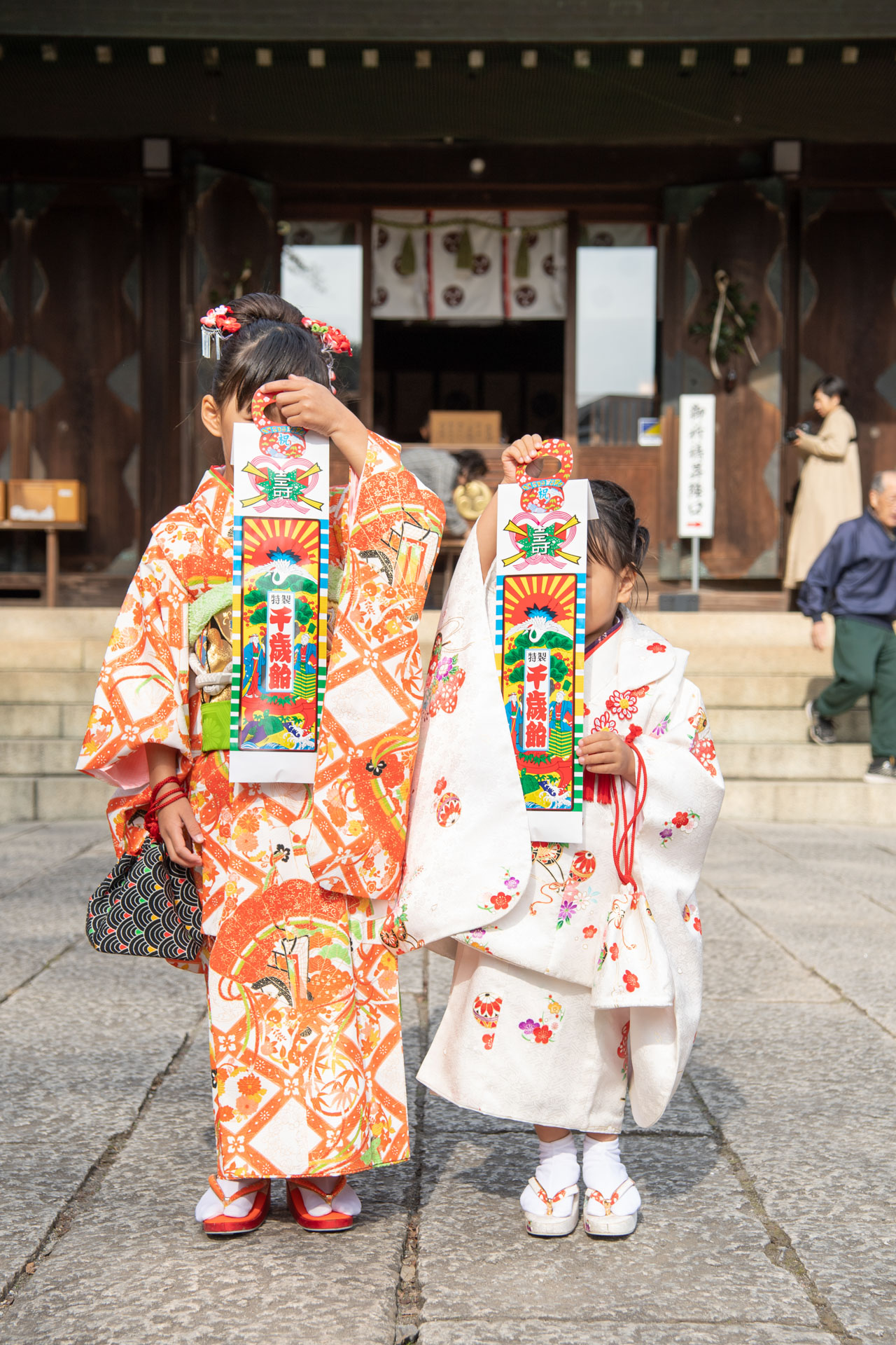 所澤神明社の七五三千歳飴の袋で顔を隠し社殿の前に立つ姉妹